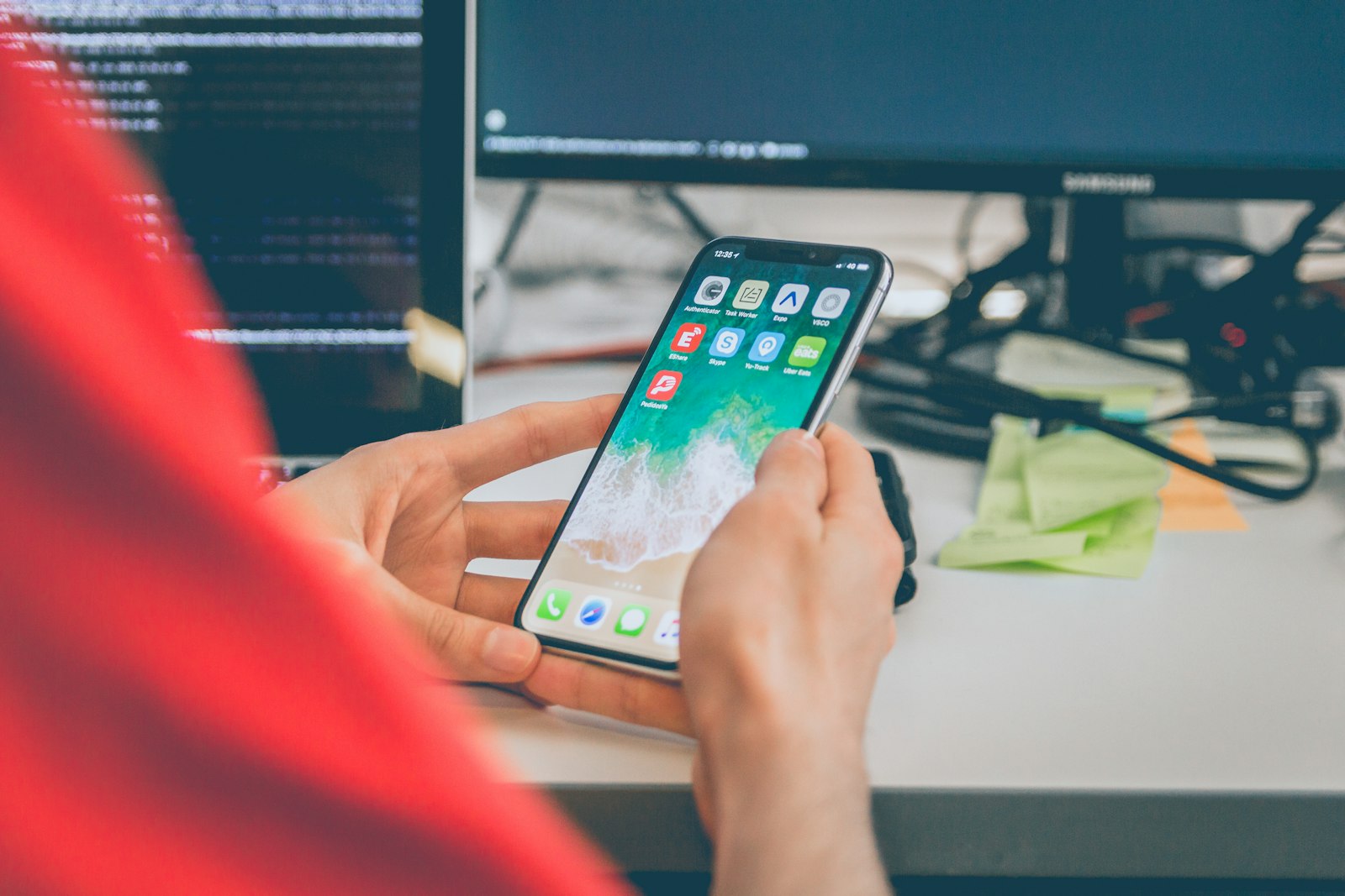 Person using a smartphone at a desk with code on the monitors behind